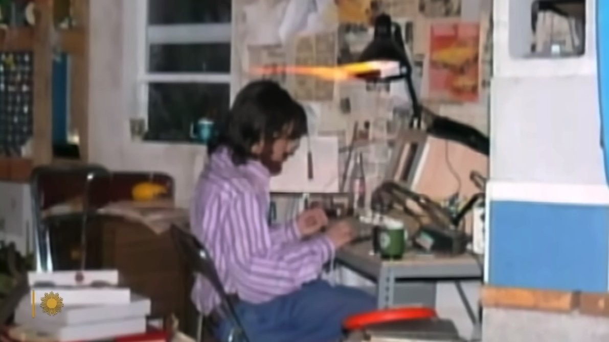 A person sits at a cluttered desk with tools, papers, and a lamp, in a room with newspaper-covered walls and a window.