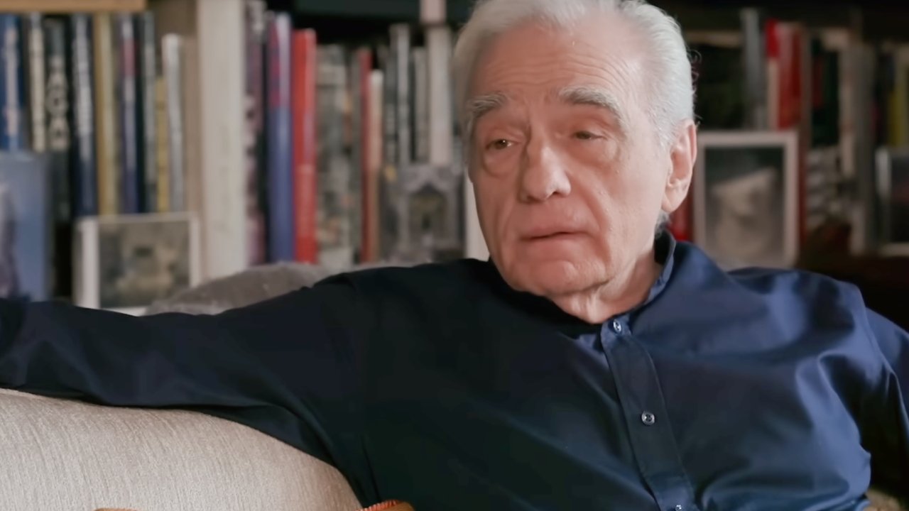 Elderly person with white hair wearing a dark shirt, sitting on a beige couch in front of a bookshelf filled with various books.