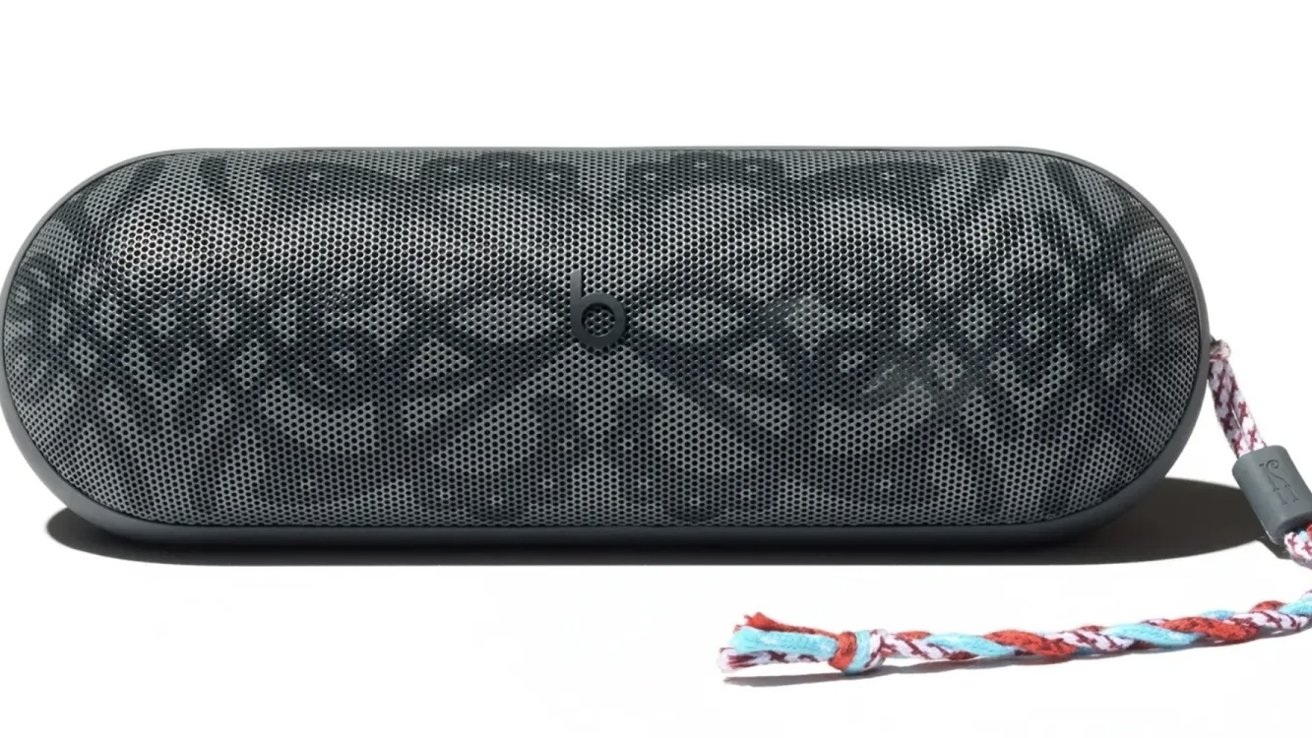 Black portable speaker with patterned front and rounded edges, featuring a woven multicolored strap attached on the right side.