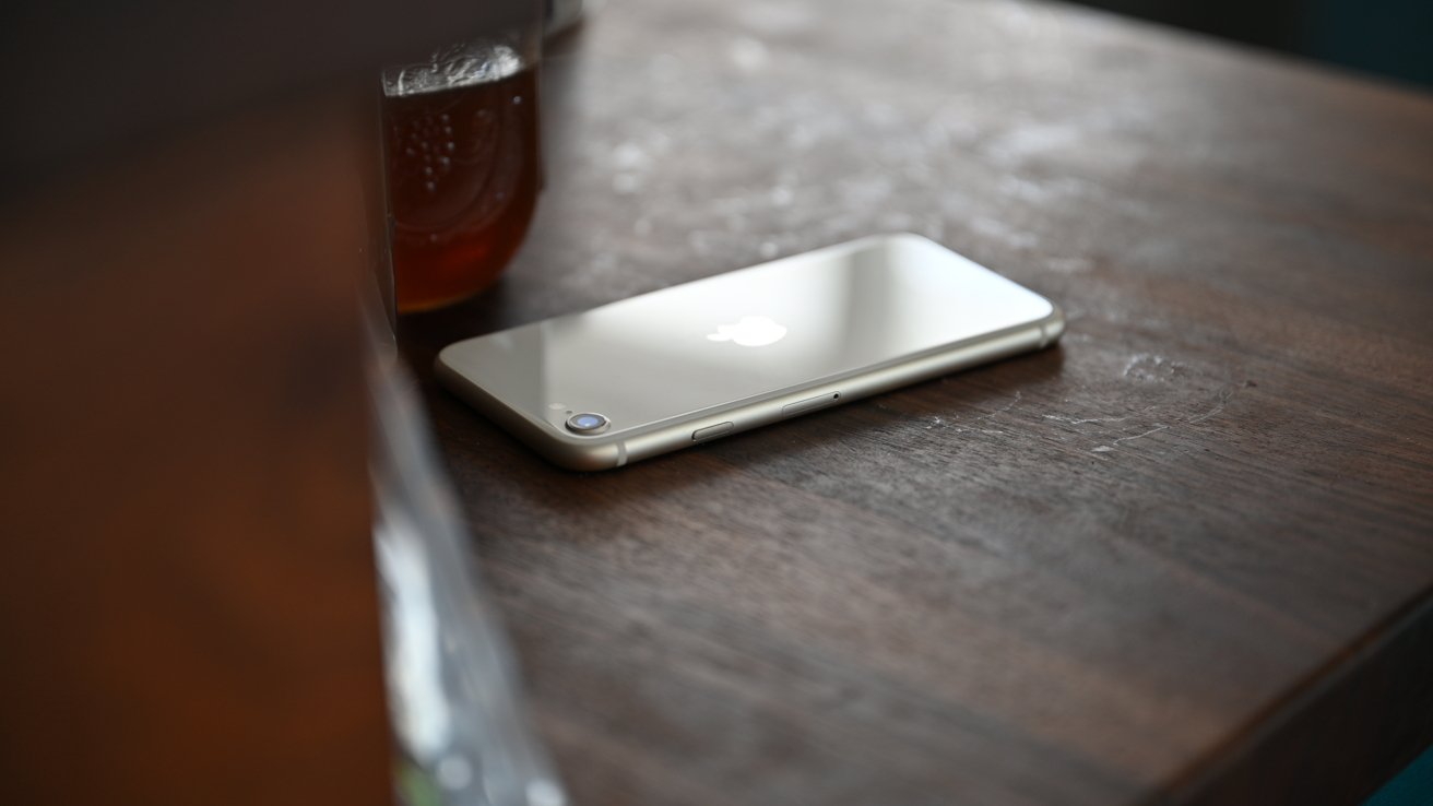 Silver smartphone with a bitten apple logo rests face down on a wooden table next to a glass with brown liquid.
