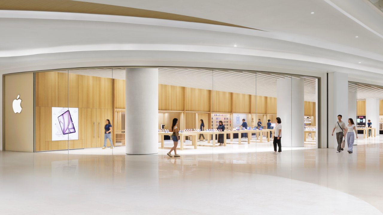 Bright, spacious retail store with wooden interior, glass walls, and people browsing products on tables. A prominent logo is displayed on one side.