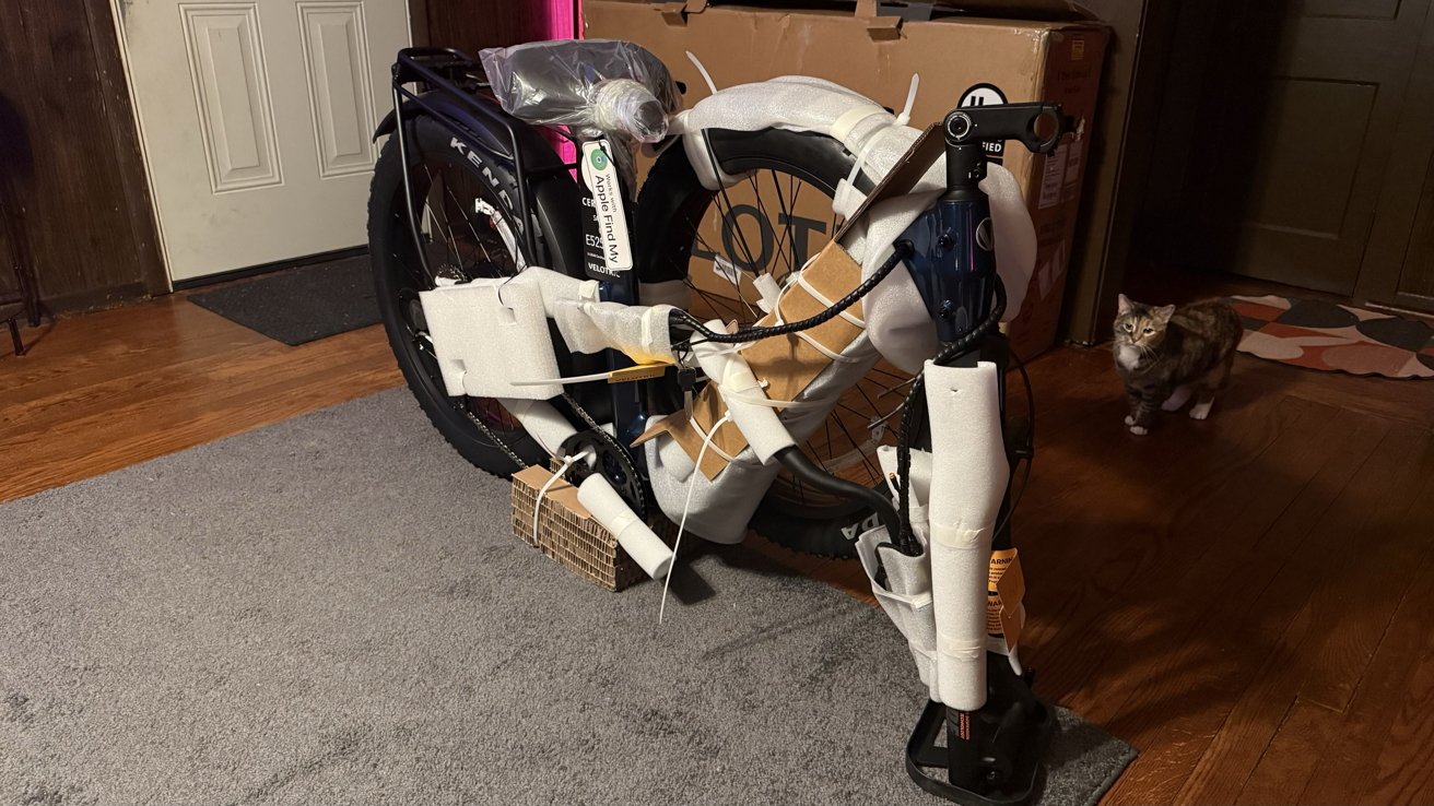 Partially assembled bicycle wrapped in protective packaging stands indoors on a rug. A cat curiously observes in the background.