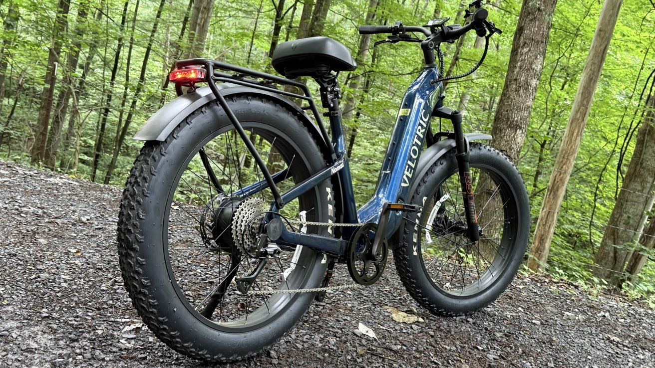 Blue electric fat-tire bike on a gravel path in a lush, green forest setting.