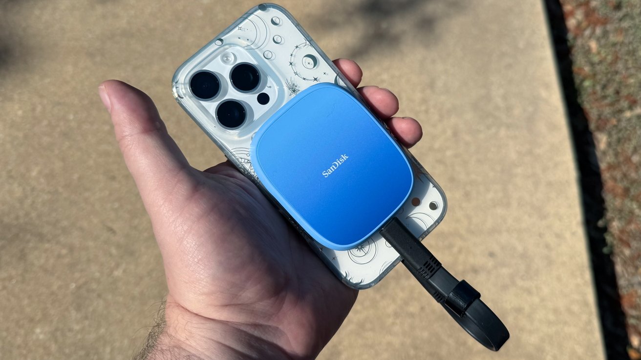 Hand holds a smartphone with a decorative case with a blue SSD portable unit with Blue Sandisk, connected by a black cable, via a concrete background.