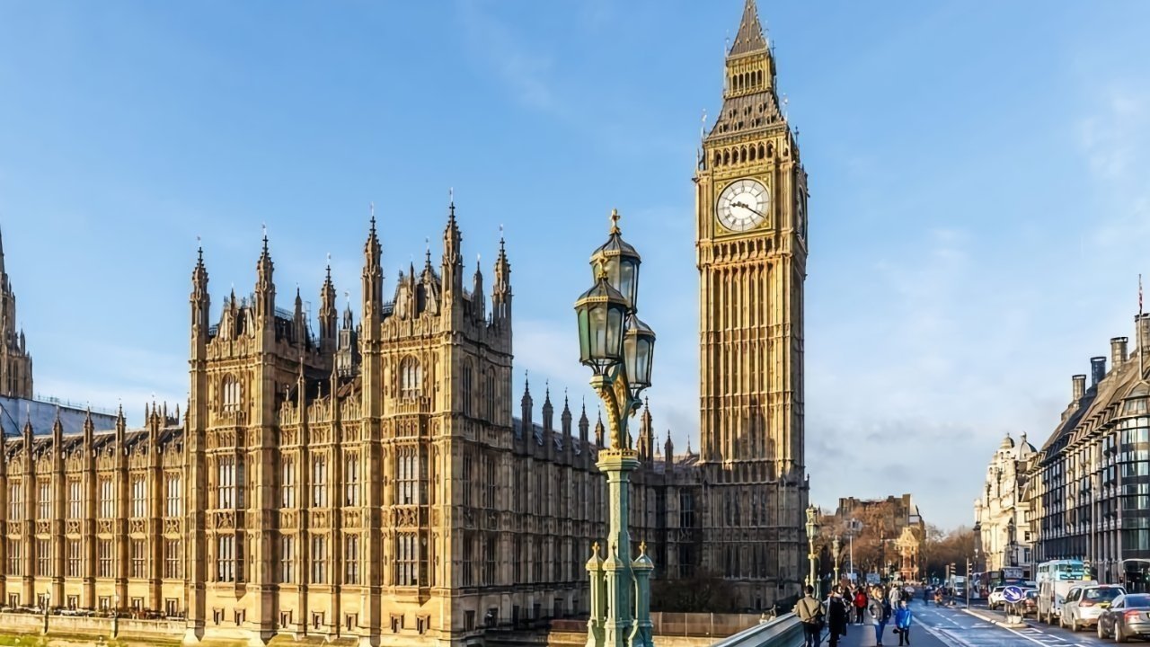 Gothic-style clock tower and parliament buildings with spires, street lamps in foreground, people walking on bridge under clear blue sky.