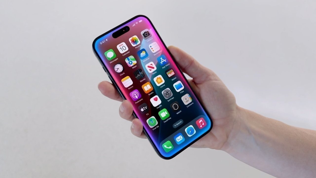 A hand holds a smartphone displaying a colorful grid of app icons against a plain, light background.