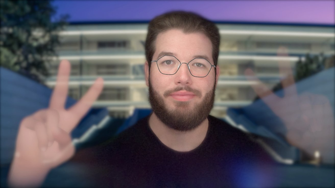 A man with glasses and beard holds up two peace signs against a blurred, modern building background with trees, under a purple and blue sky.