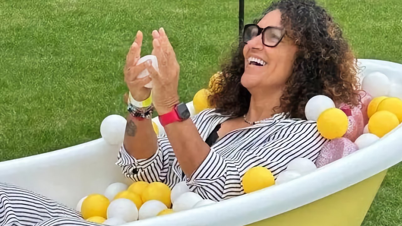 Person wearing glasses and striped clothes, laughing while sitting in a bathtub filled with white and yellow balls on a grass lawn.