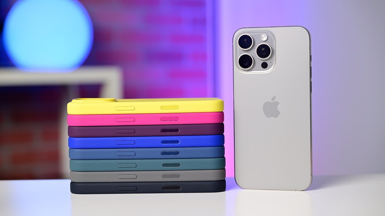 A silver smartphone with triple camera lenses stands upright next to a stack of colorful phone cases on a white surface, with a blurred blue and purple-lit background.