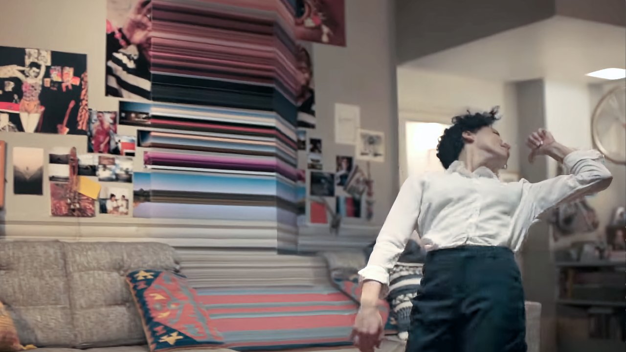 Person wearing a white shirt and dark pants dances energetically in a room with colorful wall art and photos.