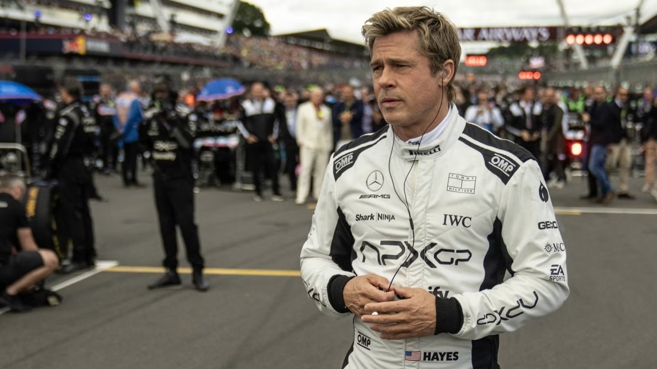 Man in a racing suit with logo patches stands on a crowded racetrack, wearing an earpiece, surrounded by people and vehicles in the background.