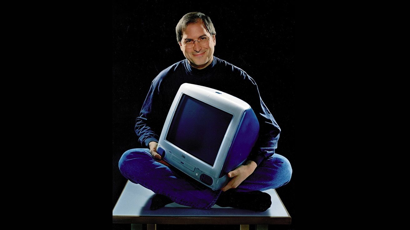 Smiling man sits cross-legged holding a vintage computer monitor, wearing blue jeans and a dark sweater, against a black background.