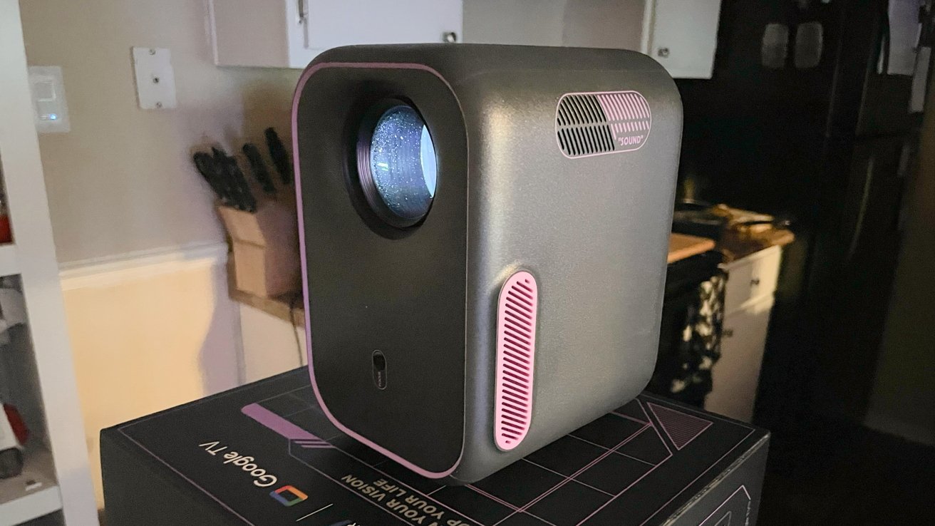 Compact gray projector with pink accents sits on a box in a dimly lit room, with kitchen in the background.