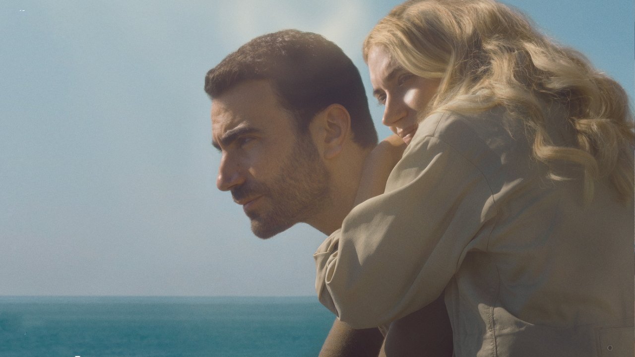 A man and woman embrace while gazing thoughtfully out at the ocean under a clear blue sky.