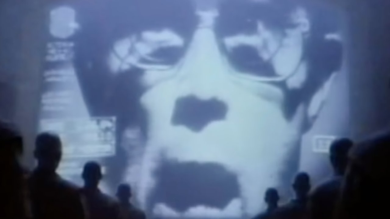 Large face on screen wearing glasses, speaking to a crowd of silhouetted figures in a dark room.
