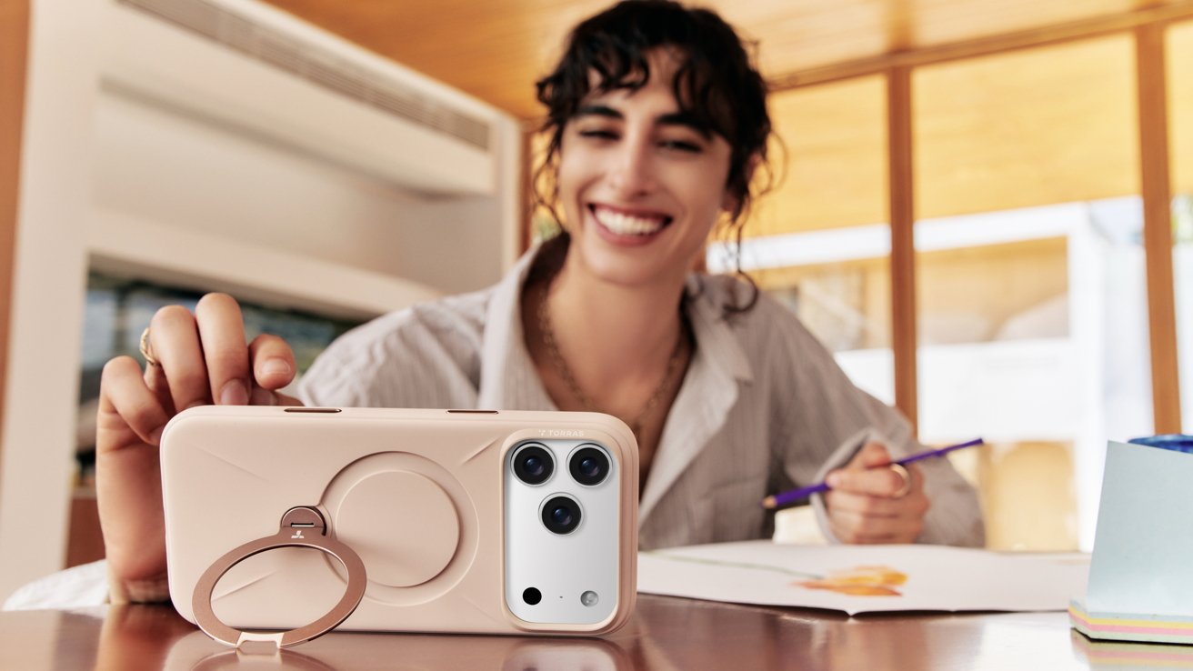 Smiling person holding smartphone in a case with a circular stand, drawing on paper with a pencil, indoors with wooden decor.