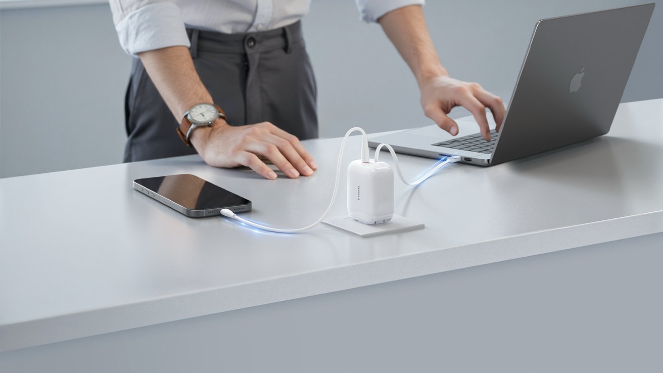 Person standing at a desk using a laptop, with a smartphone connected to a charger emitting light, both devices charging simultaneously.