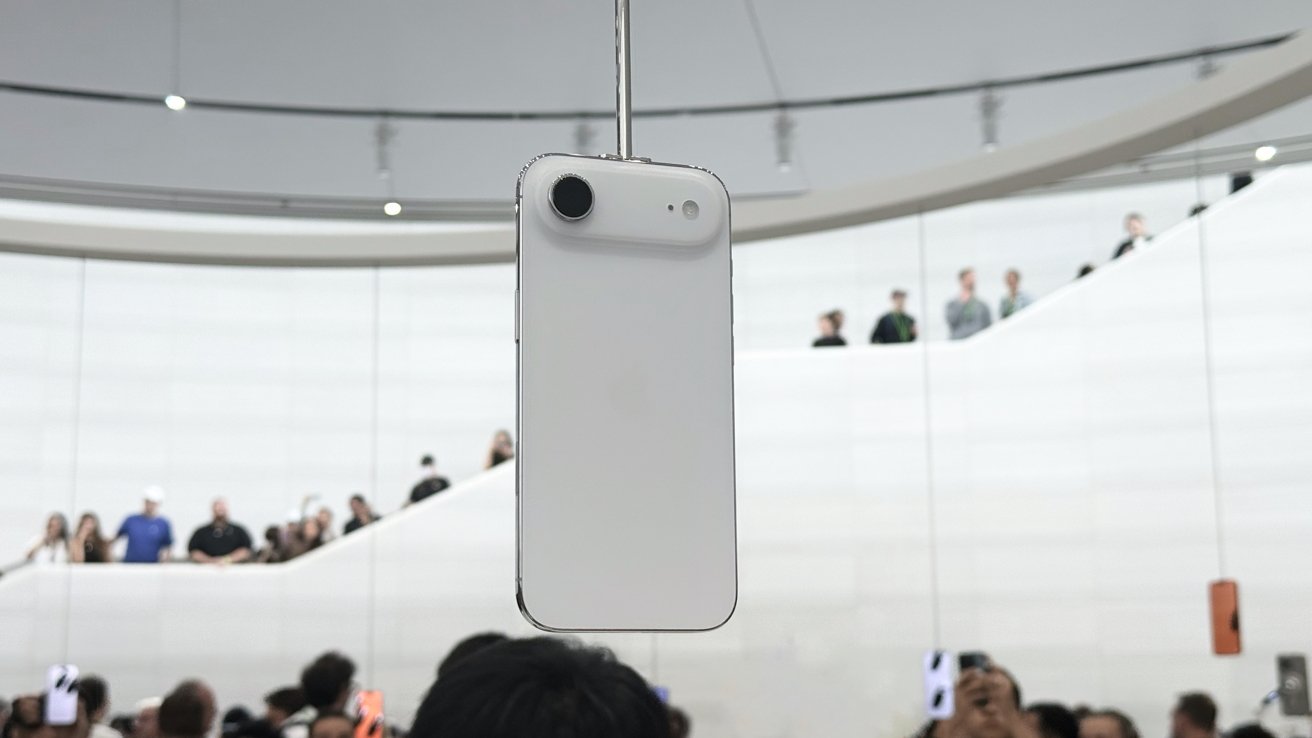 White smartphone suspended in front of a curved, modern staircase with a crowd of people in the background.