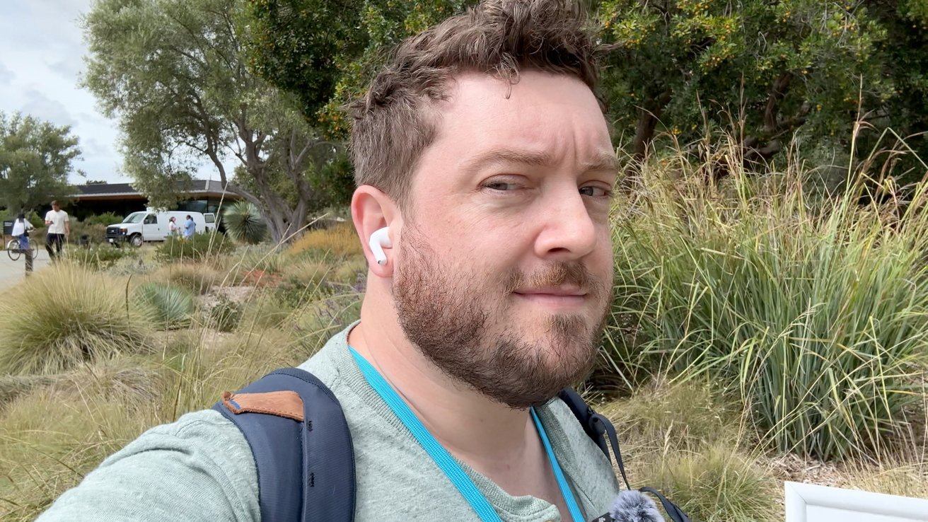 Man with curly hair and a beard wearing earbuds and a backpack, standing outdoors with grass and trees in the background.