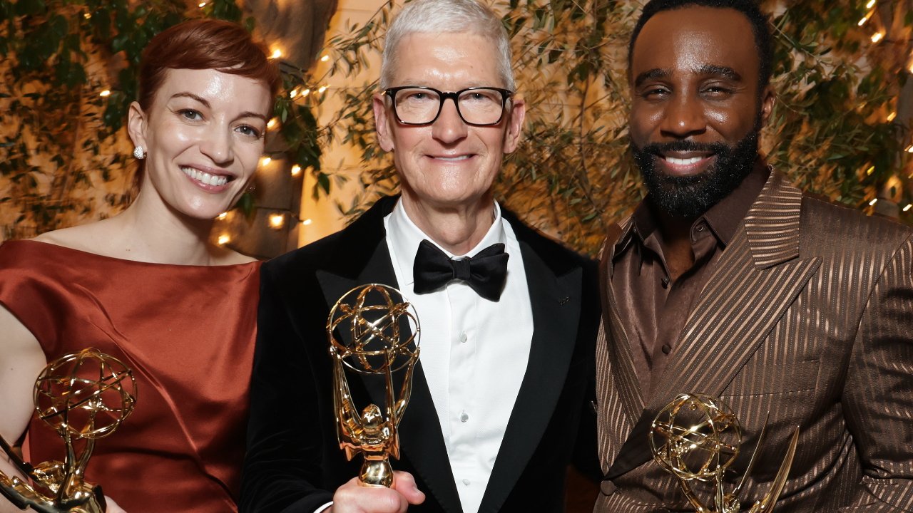 Three smiling people holding awards, dressed formally with elegant lighting and greenery in the background.