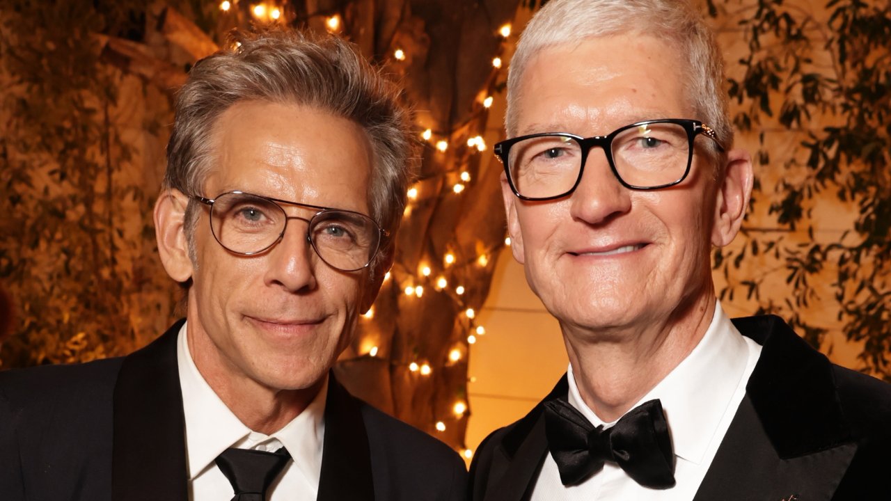 Two men in suits and glasses smile, standing in front of a warmly lit, leafy backdrop.