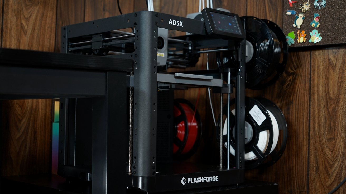 A 3D printer, labeled Flashforge AD5X, with spools of filament on a wooden table beside colorful magnets on a corkboard.