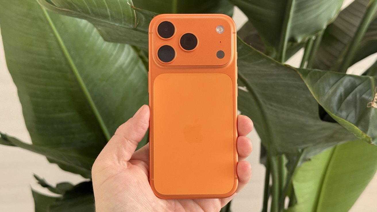 A hand holds an orange smartphone with three camera lenses, in front of large green leaves.