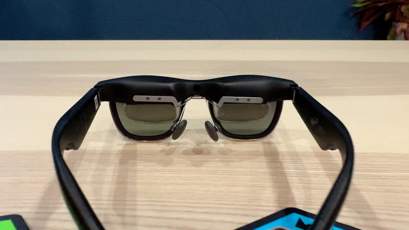 Black intelligent glasses with black reality with strong frames resting on wooden surfaces, small metal details and an integrated camera between lenses.