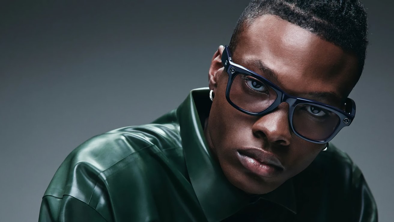 A person with short hair wearing large glasses and a shiny dark green jacket and enjoys forward with a serious expression against a gray background.