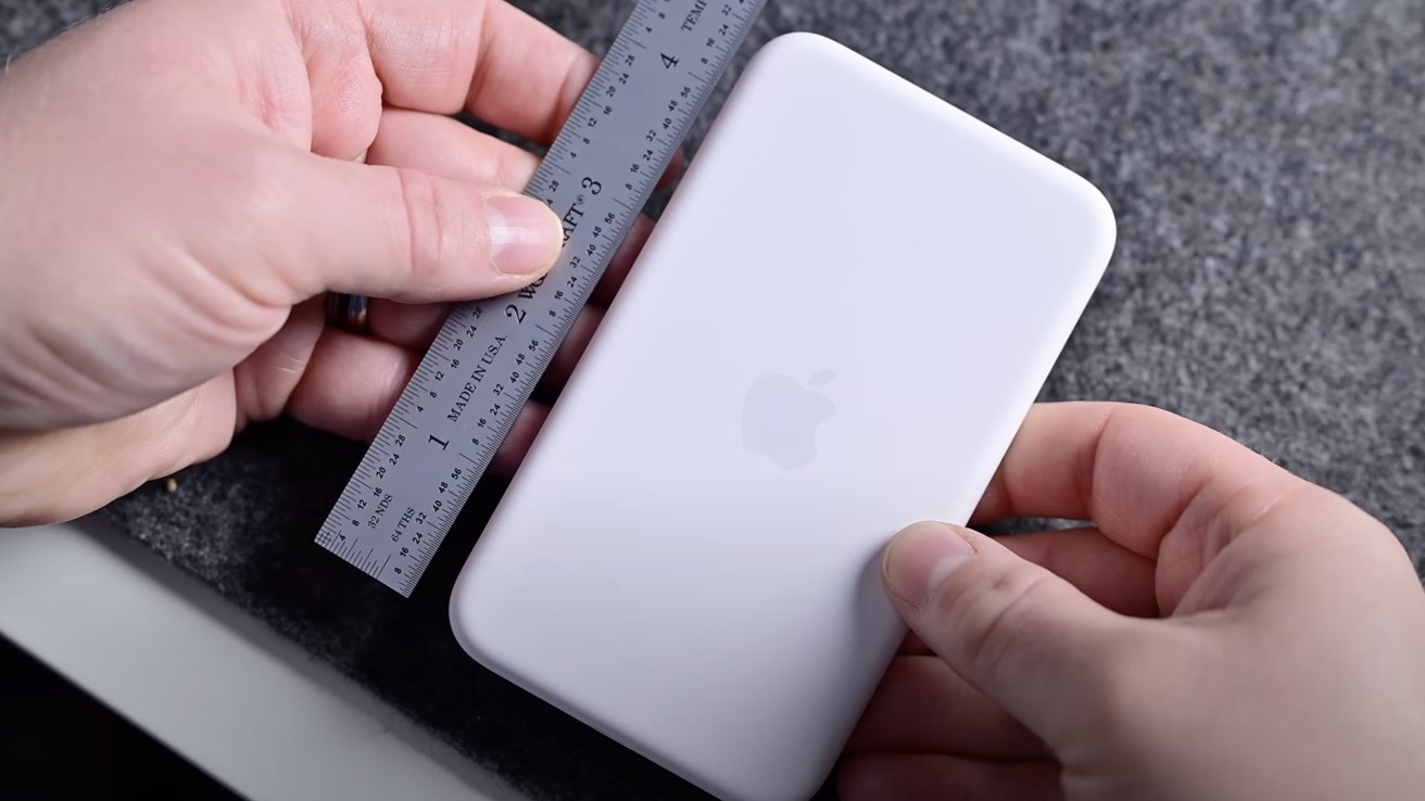 Hands holding a small white rectangular device with rounded corners, featuring a faint Apple logo, alongside a metal ruler measuring its length.