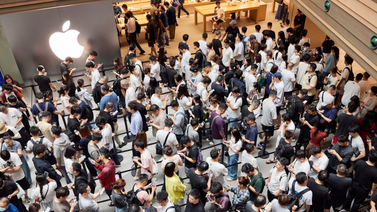 Crowded store with many people lined up, holding smartphones, beneath a large Apple logo, in a modern retail environment with wooden tables.