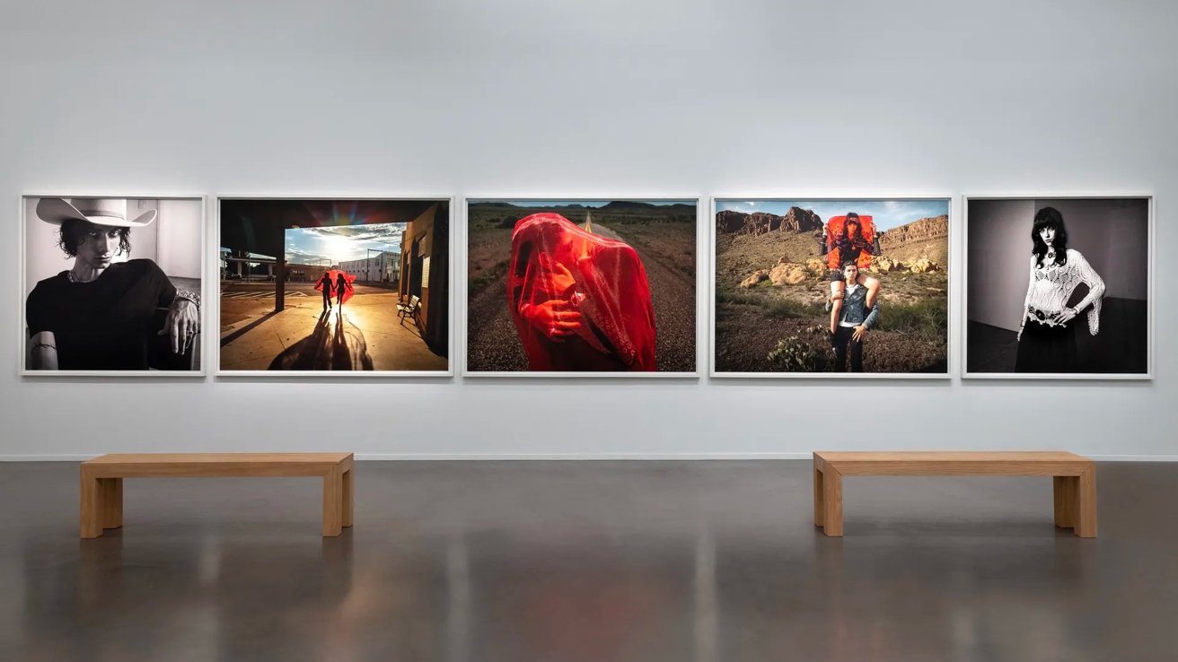 Five large framed photographs line a white gallery wall above two wooden benches. The images include portraits and outdoor scenes featuring people in various settings.