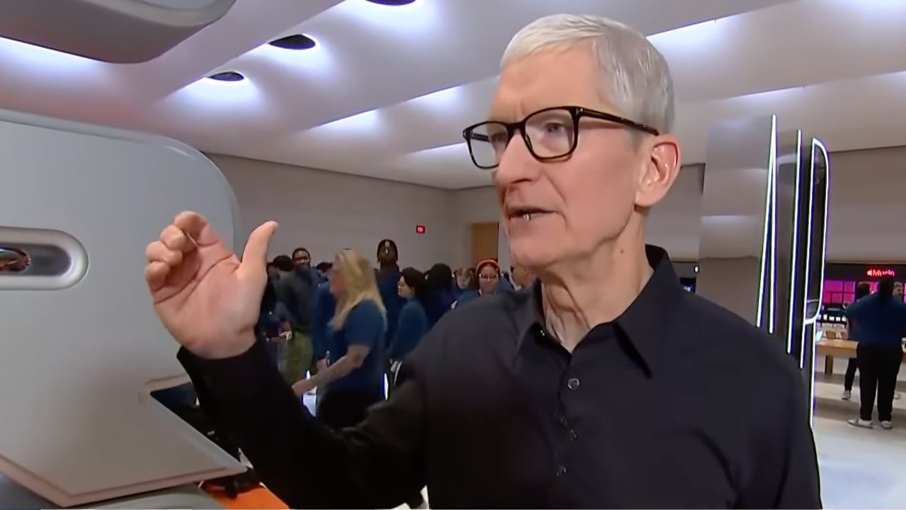 Person with gray hair and glasses gestures while speaking in a modern, well-lit store. Background features people standing and sleek architectural elements.