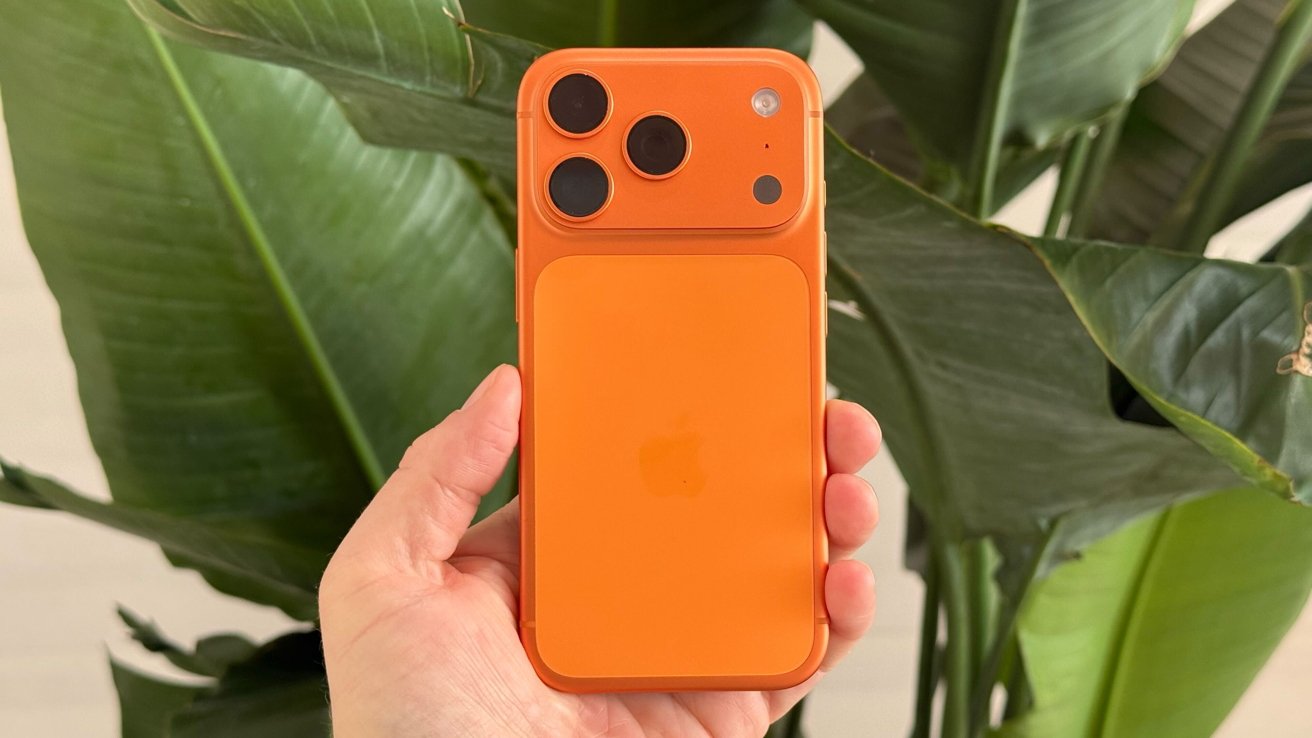 Orange smartphone with three camera lenses held in hand, leafy green plant background.