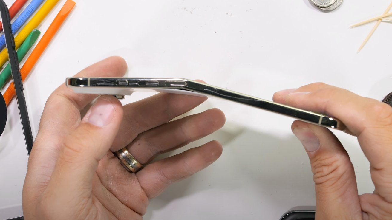 Hands holding a bent smartphone on a white surface, with colorful pencils, a coin, and toothpicks in the background.