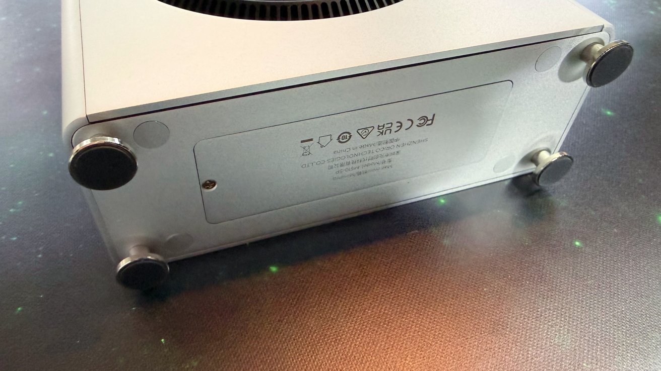 Silver rectangular electronic device base with four round feet. Engraved symbols and text visible on panel. Placed on a dark surface with faint green spots.