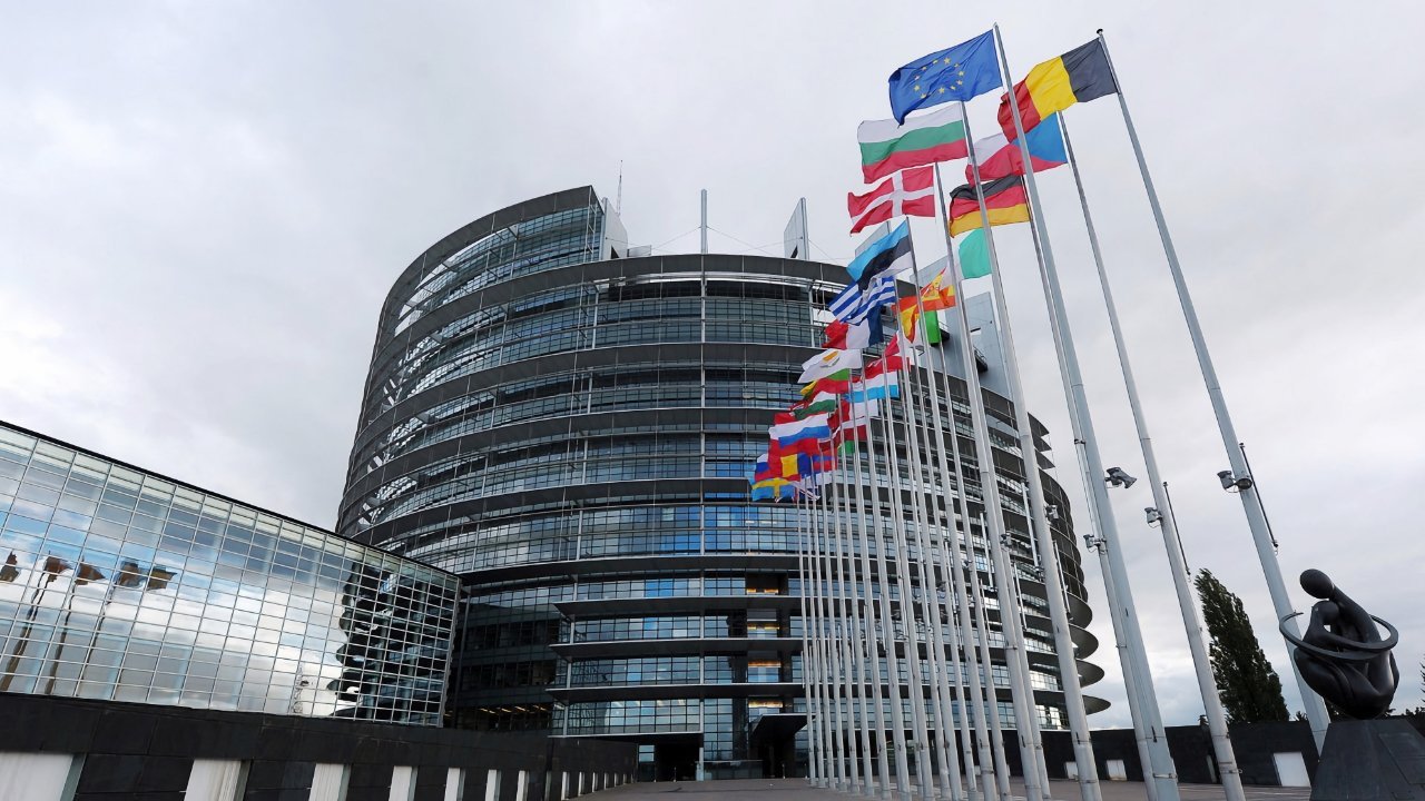 Curved glass modern building with numerous international flags on poles and a sculpture in the foreground under a cloudy sky.