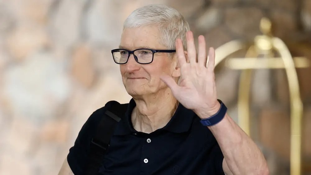 Man with short white hair, wearing glasses and a dark polo shirt, waves with right hand raised in front of a blurred neutral background.