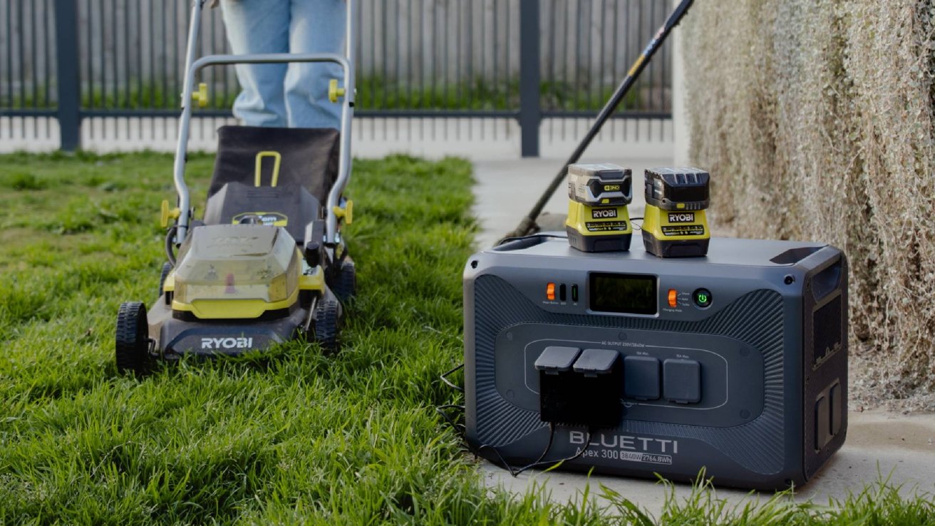 Person with a lawnmower on grass beside a portable power station with two batteries on top.