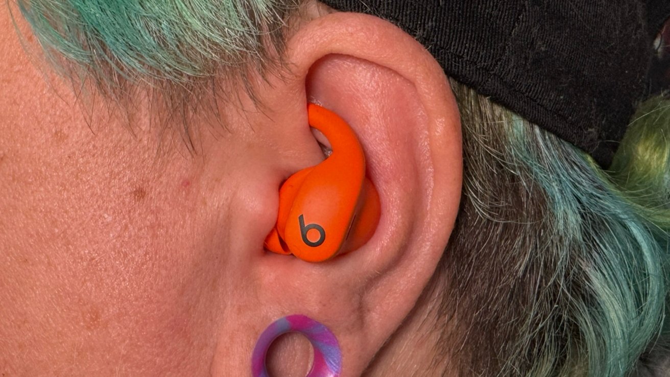 A person with teal hair wears orange earbuds in their ear, featuring a stretched earlobe with a colorful plug.