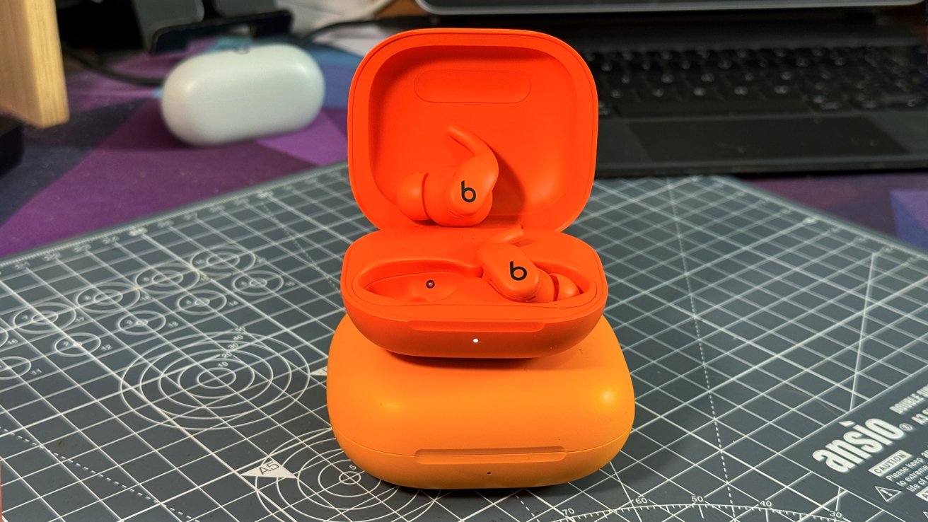 Orange wireless earbuds with black 'b' logo in open charging case, placed on another closed case, on a grid-patterned surface.