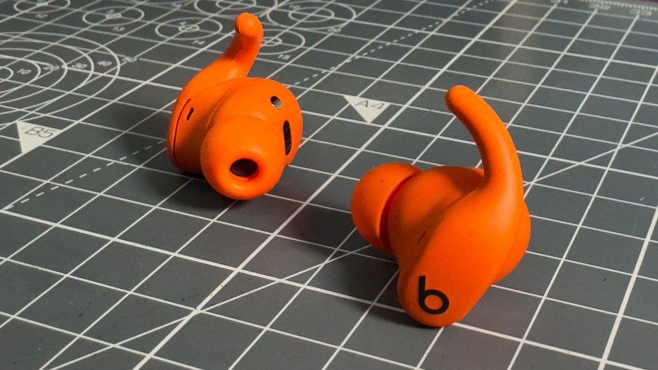 Two bright orange wireless earbuds with ear hooks on a gray grid-patterned surface.