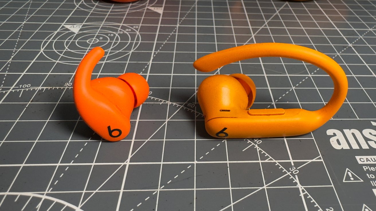 Orange wireless earbuds with ear hooks on a gray, grid-patterned cutting mat.