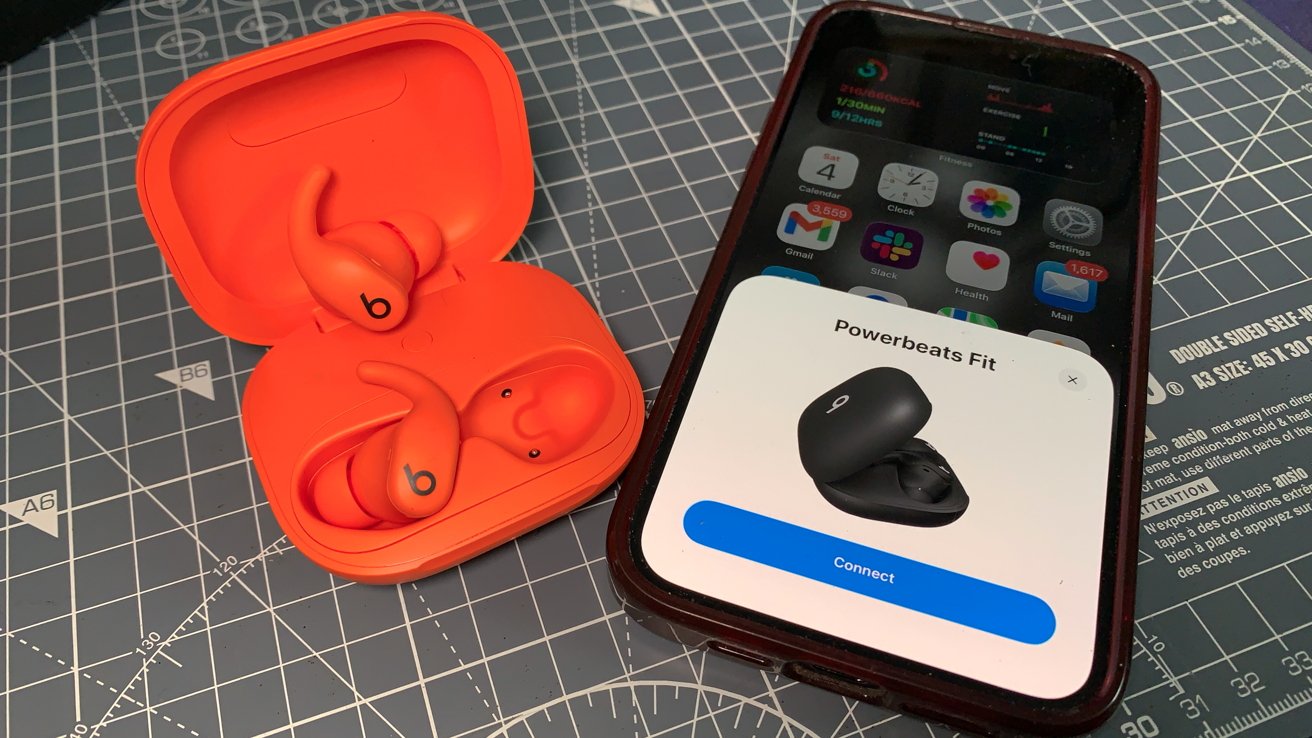 Orange wireless earbuds in an open case next to a smartphone displaying a Powerbeats Fit connection screen, on a cutting mat.