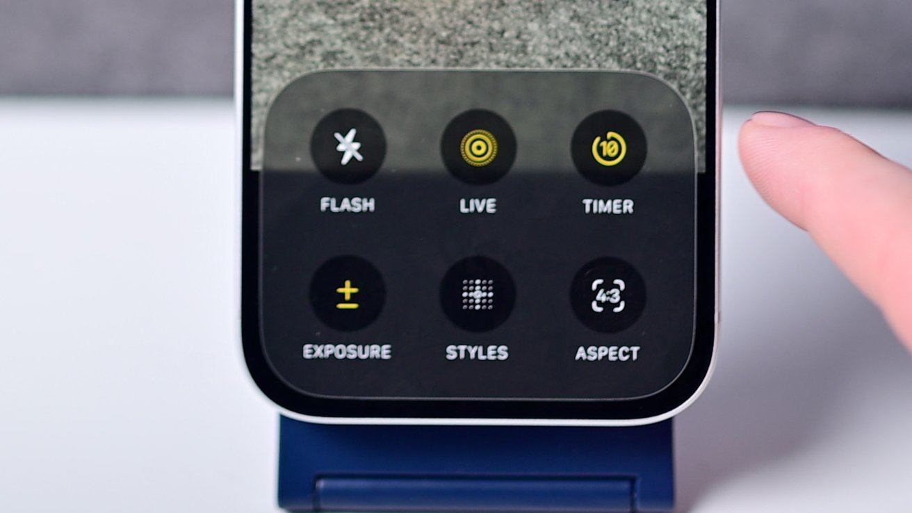Smartphone camera settings screen showing options for flash, live, timer, exposure, styles, and aspect ratio; a finger hovers over the screen.