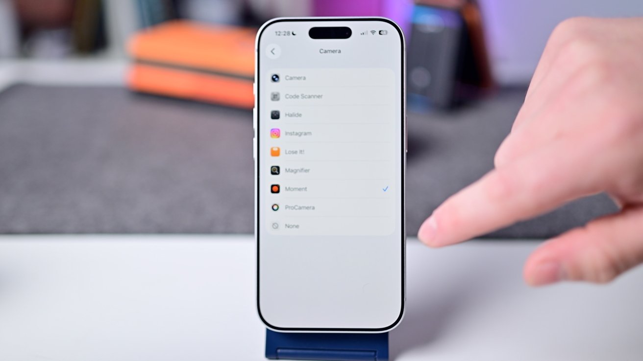 Smartphone displaying an app selection menu with a finger pointing at the screen. Blurred background with books and objects.