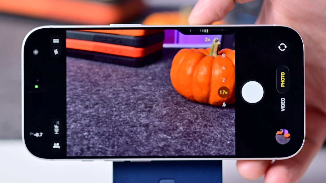 Phone displaying a photo preview of a small orange pumpkin on a gray surface, with a hand holding the device.