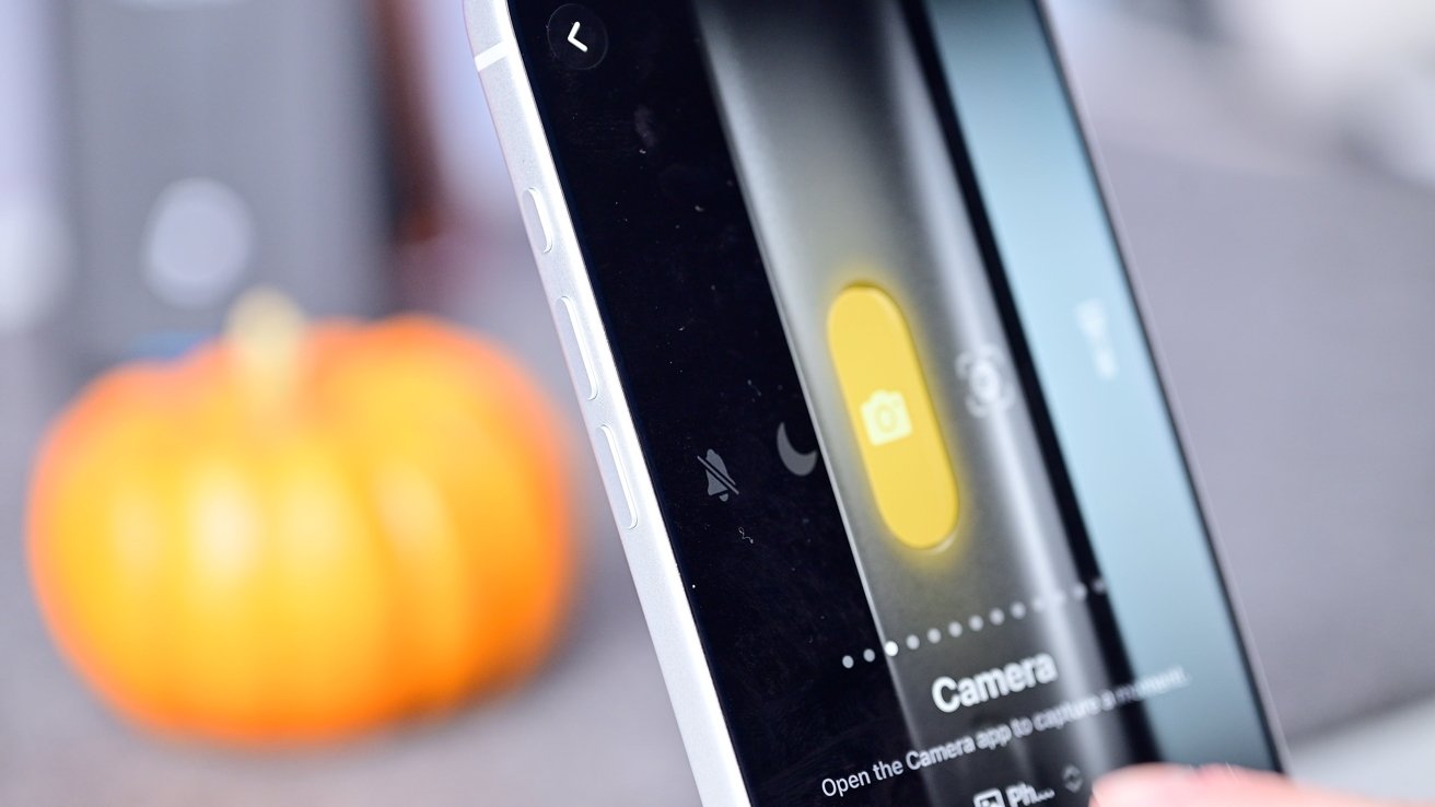 Close-up of a smartphone screen showing camera controls, with a blurred orange pumpkin in the background.