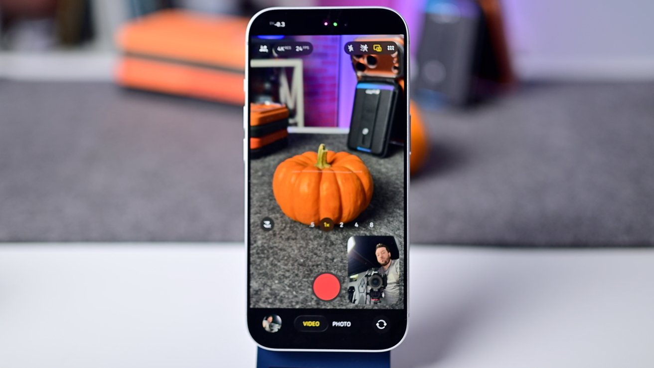 Smartphone displaying camera app, focusing on a small orange pumpkin on a table. Background includes blurred office items, emphasizing the pumpkin's sharpness on the screen.