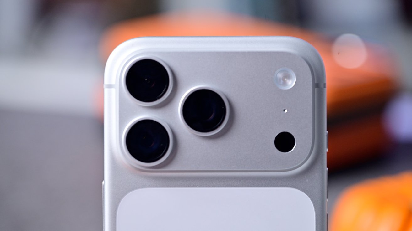 Close-up of a silver smartphone's rear camera setup with three lenses and a flash, against a blurred background.